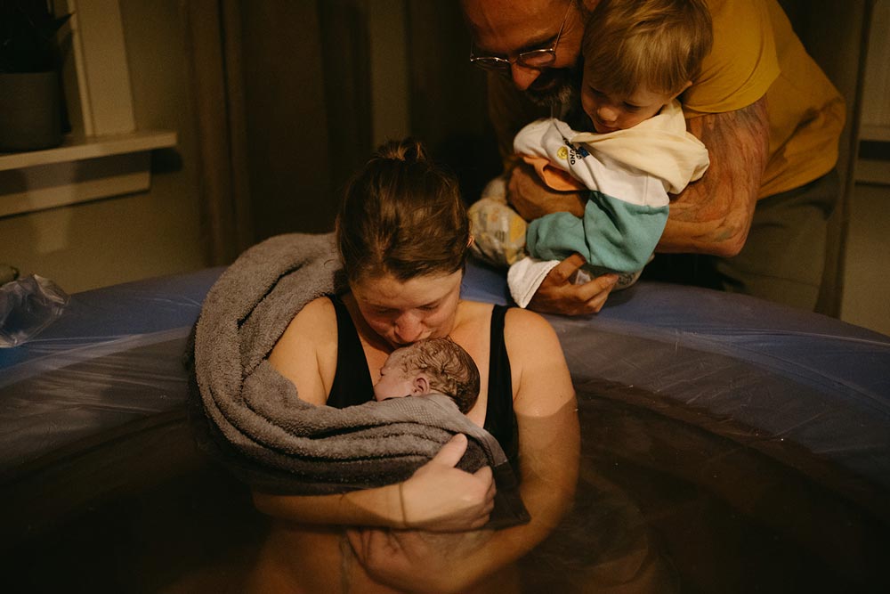 Mamma håller om sitt nyfödda barn i en födselpool medan partner och barnets syskon står glada på sidan